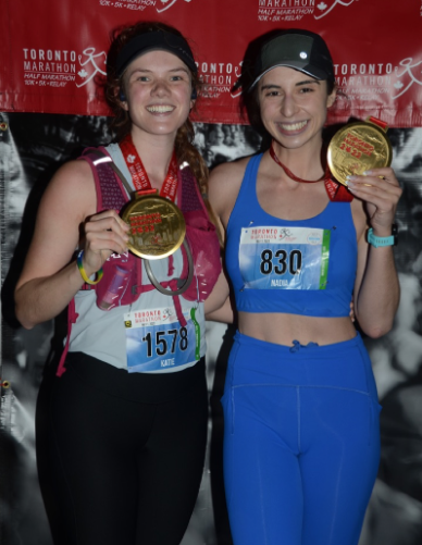 Two women wearing running gear are standing next to each other. They are smiling and holding up their medals, received after completing the Toronto Marathon. A red banner with the marathon logo on it is behind them.